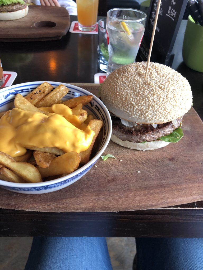 Holzbrett mit einer Schale Pommes mit Käsesauce und einem Burger, im Hintergrund ein Glas und ein weiteres Brett